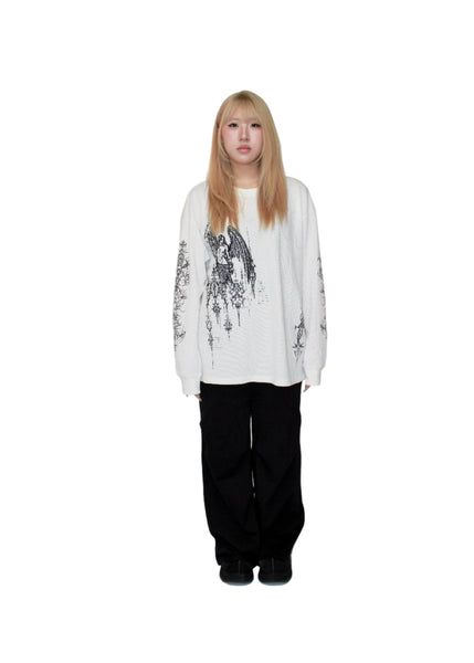 Person wearing a white cardigan with black floral patterns over a white shirt and black pants on a plain background